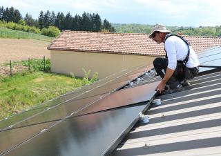 Une homme coiffé d'un bob est en train d'installer des panneaux solaires sur le toit d'un bâtiment. Autour, un paysage verdoyant.