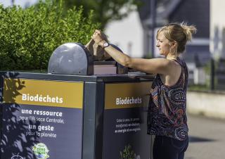 Une femme tient un sac en papier au dessus d'une cuve sur laquelle on peut lire "biodéchets, une ressource pour l'Alsace centrale, un geste pour la planète"